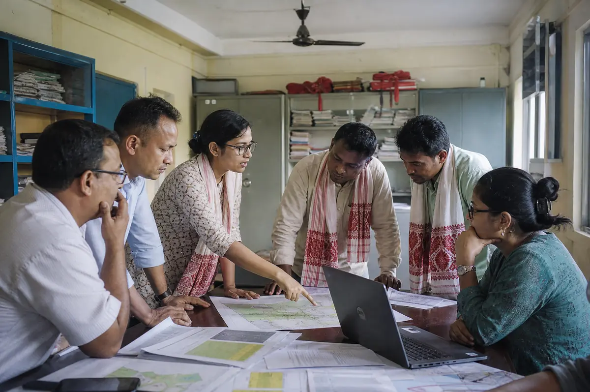 A consultant points to a village map on a table covered with district maps while block officials, an NGO manager and FPO leaders discuss a plan.